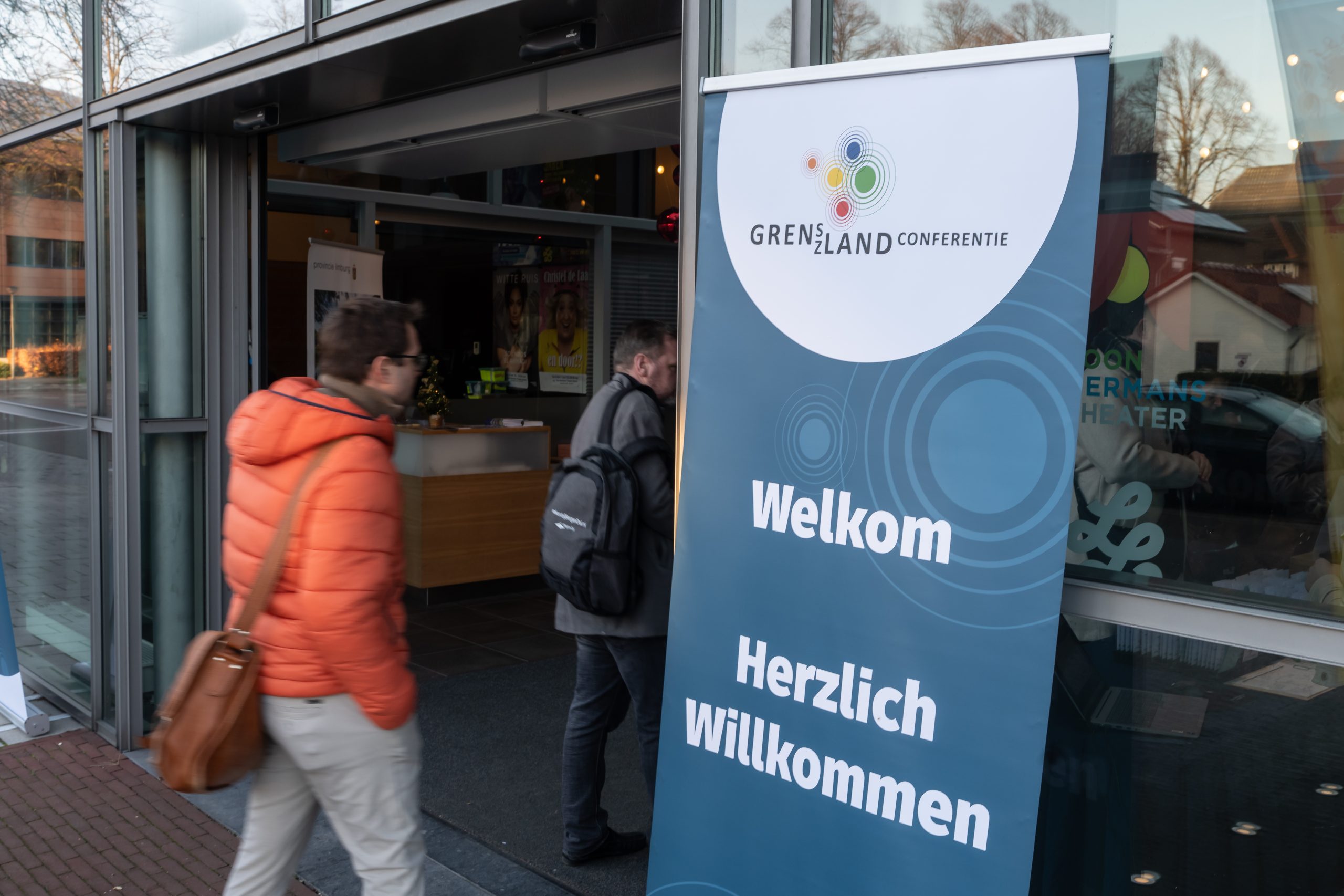 Mensen die naar binnen lopen bij de Grenslandcoferentie. Er staat een bord 'Welkom bij de Grenslandconferentie'.